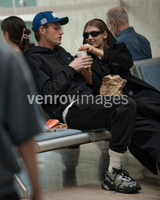 Two adults at an airport sit on a bench with a drink and snack. The woman wears sunglasses and a long coat, and the man a baseball cap.