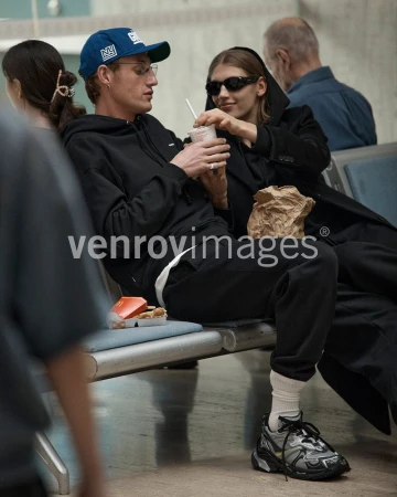 Two adults at an airport sit on a bench with a drink and snack. The woman wears sunglasses and a long coat, and the man a baseball cap.