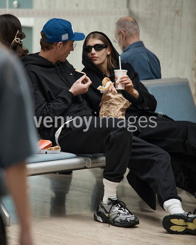 Young adult couple sits on bench and eats takeout. The woman wears sunglasses and a hood. Neutral indoor light.