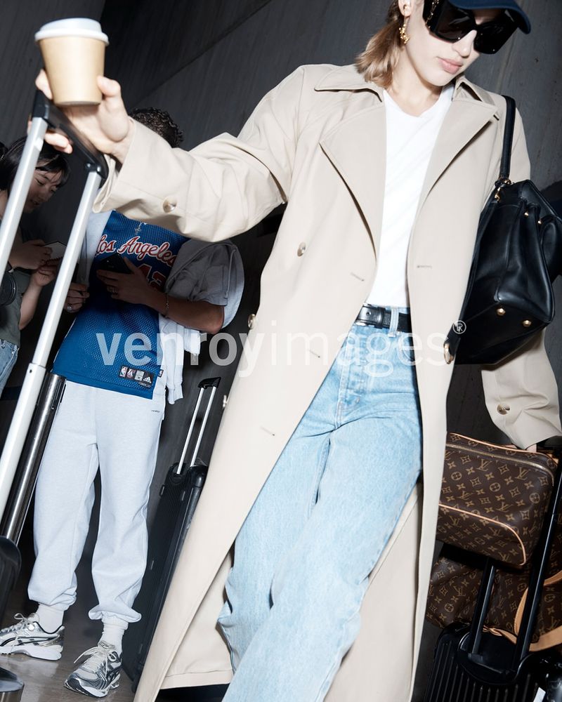 A woman in a trench coat walks through a modern building with luggage, carrying coffee, styled in fashionable clothes with people in background.