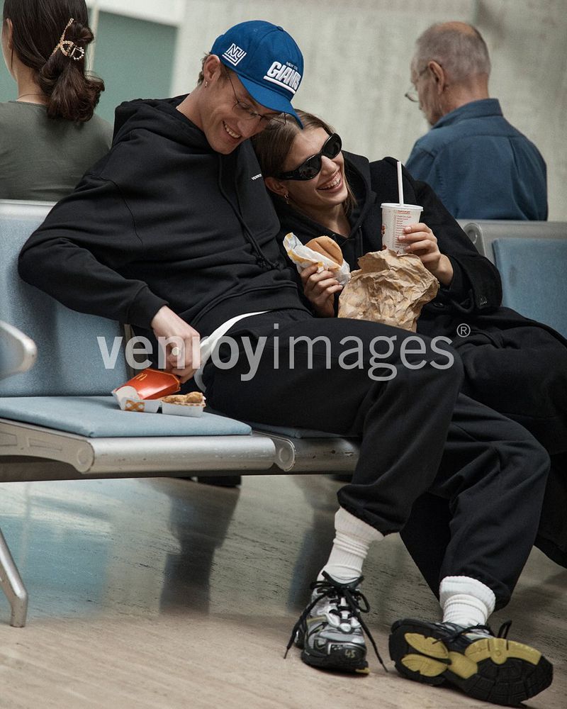 Two people, an adult male and adult female, sit on a bench eating fast food; the man wears a blue baseball cap and the woman wears sunglasses.