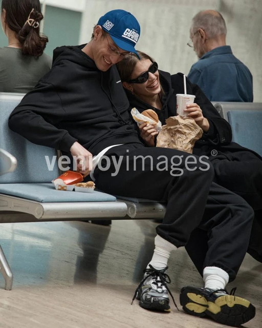 Two people, an adult male and adult female, sit on a bench eating fast food; the man wears a blue baseball cap and the woman wears sunglasses.