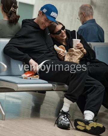 Two people, an adult male and adult female, sit on a bench eating fast food; the man wears a blue baseball cap and the woman wears sunglasses.