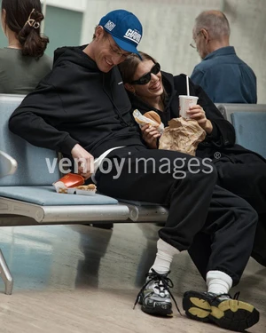 Man in black hoodie and blue cap sitting next to a smiling woman holding a burger and drink in an airport terminal.
