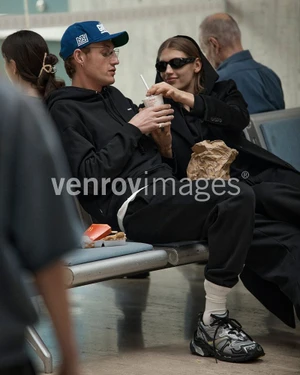 Man and woman seated on airport or station seating sharing a drink with fast food and luggage paper bag