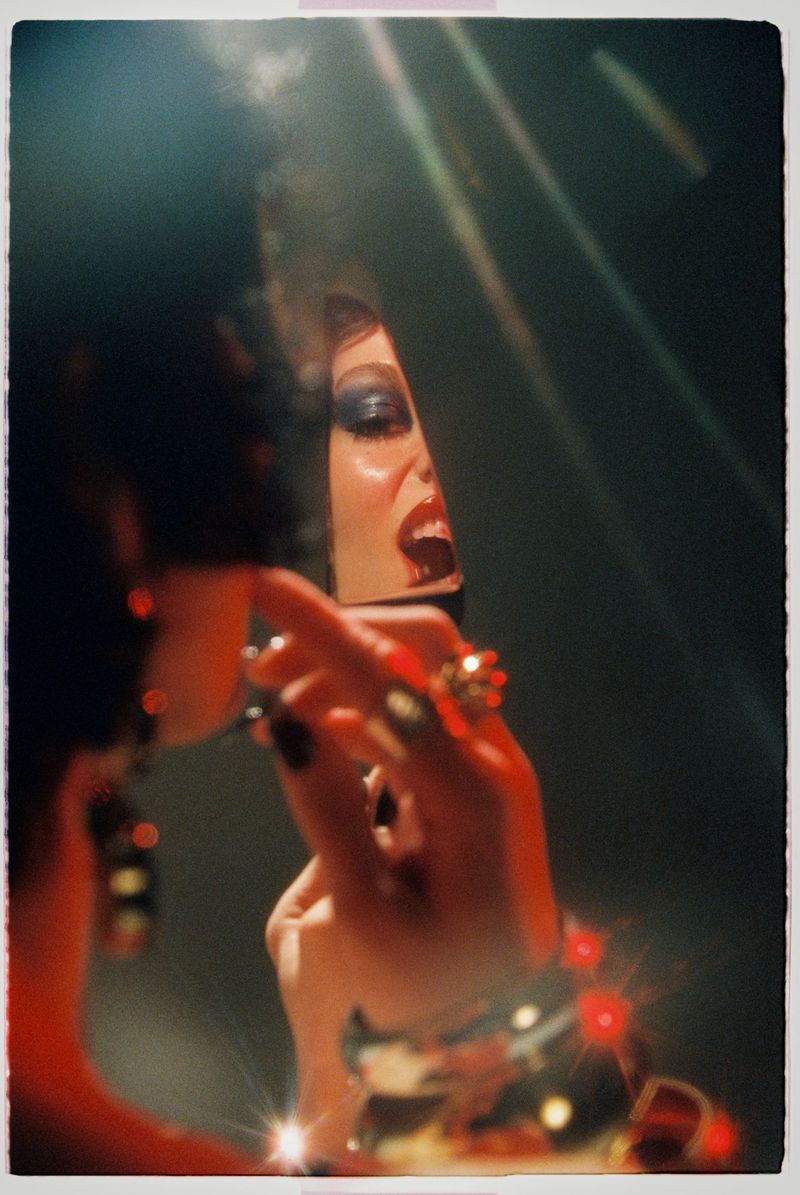 Close-up of a woman applying lipstick while looking into a mirror in a dramatic, dark setting with light flares.