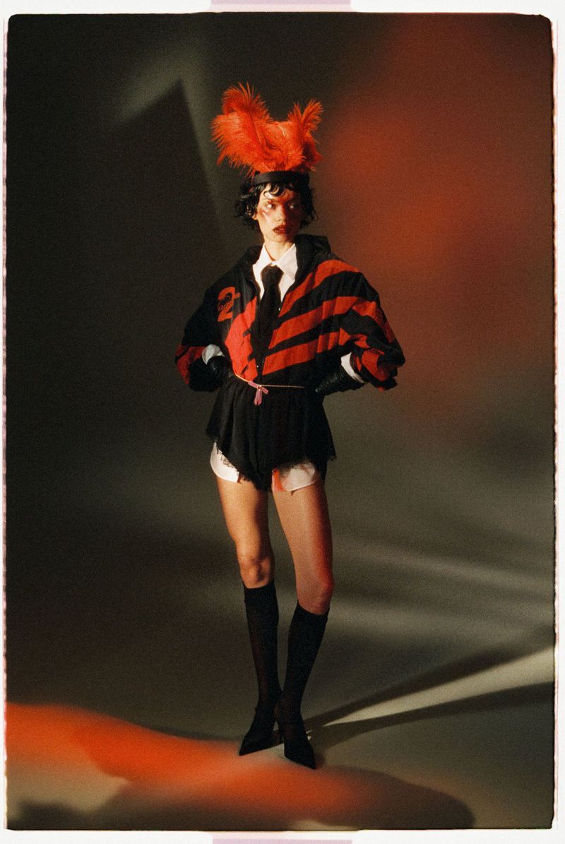 Model in a red and black striped jacket with an orange feathered headpiece posing against a gradient studio background.