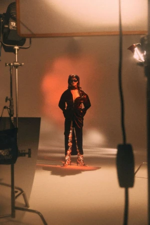 Model standing in a studio with lighting equipment framing the shot against a seamless background with warm lighting.