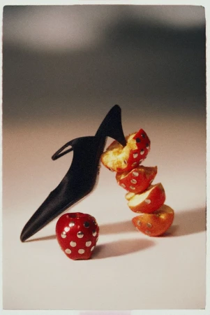 A black stiletto shoe crushing sliced red apples studded with silver hardware against a neutral studio backdrop.