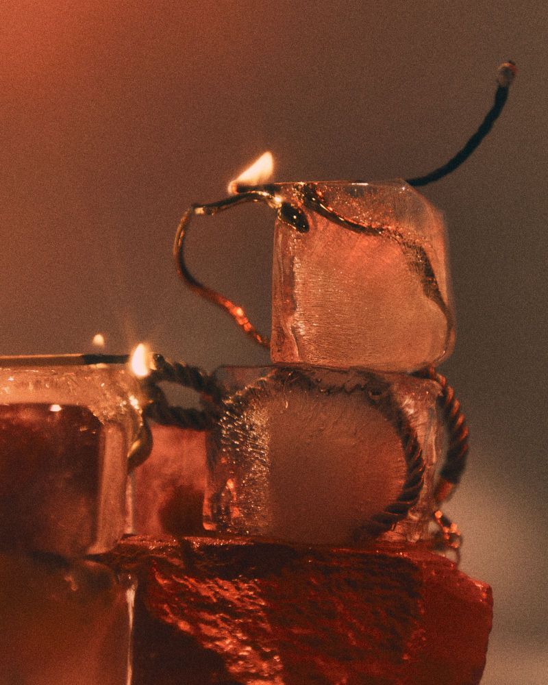 Close-up of stacked ice cubes wrapped in smoldering rope and lit matches with a warm orange glow against a neutral background.