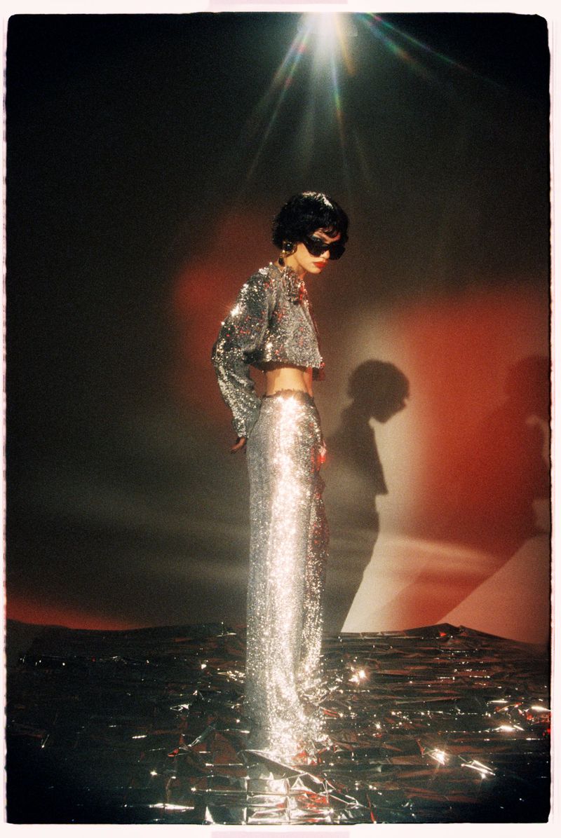 Model in shiny silver sequined two-piece outfit and dark sunglasses standing on a reflective surface under a bright overhead spotlight