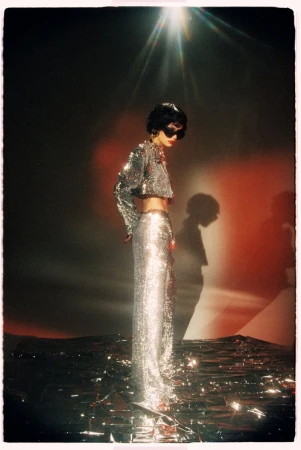 Model in shiny silver sequined two-piece outfit and dark sunglasses standing on a reflective surface under a bright overhead spotlight