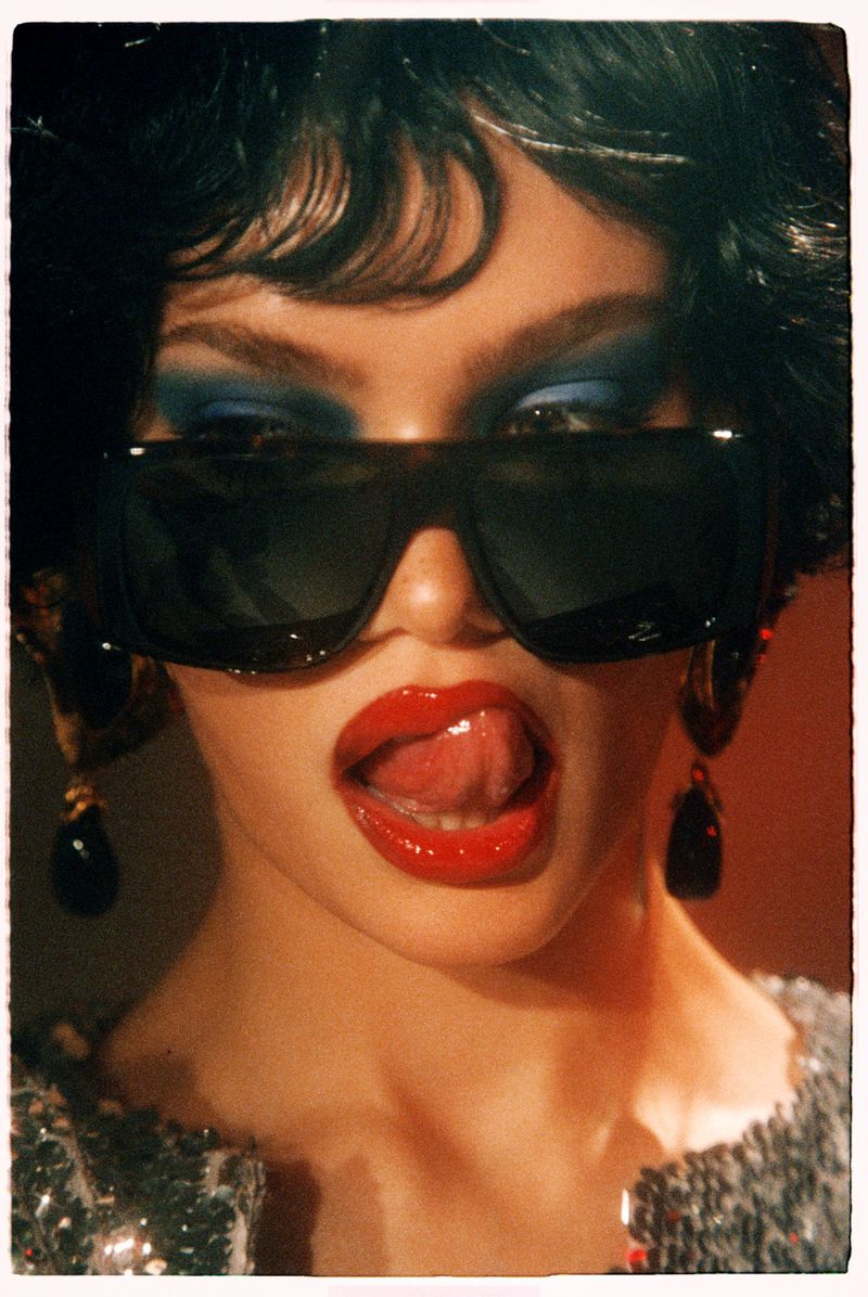 Close-up portrait of a woman wearing dark sunglasses and bold blue eyeshadow sticking out her tongue for Numero Netherlands