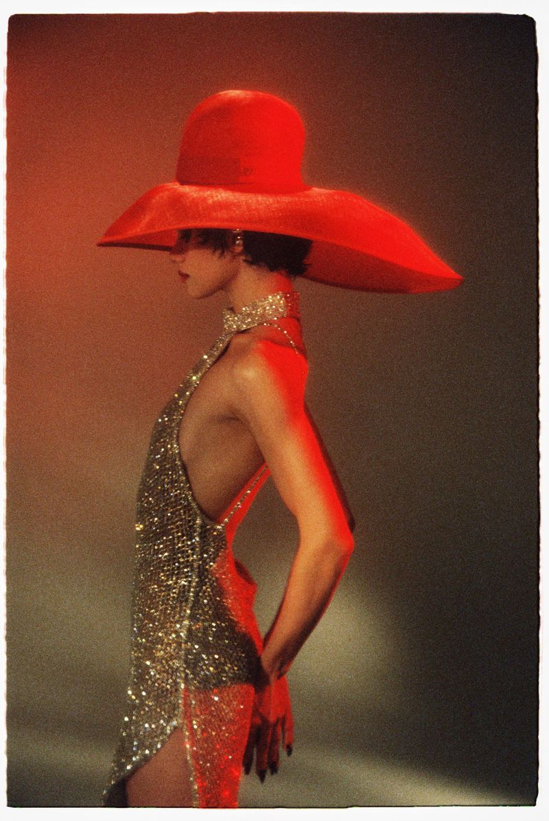 Model in profile wearing a wide-brim red hat and glittering sequin dress against a warm gradient background.