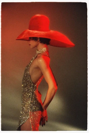 Model in profile wearing a wide-brim red hat and glittering sequin dress against a warm gradient background.