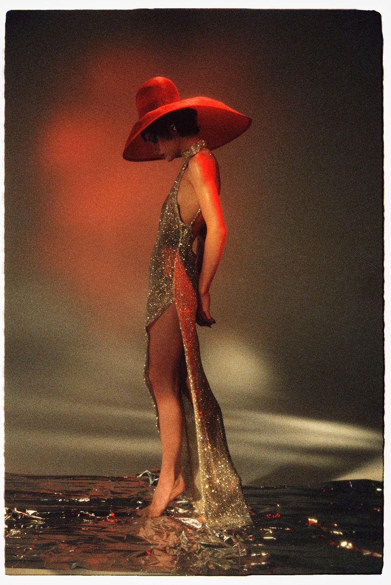 Model in a sequined gown and large red hat standing on a reflective metallic surface with warm red lighting.