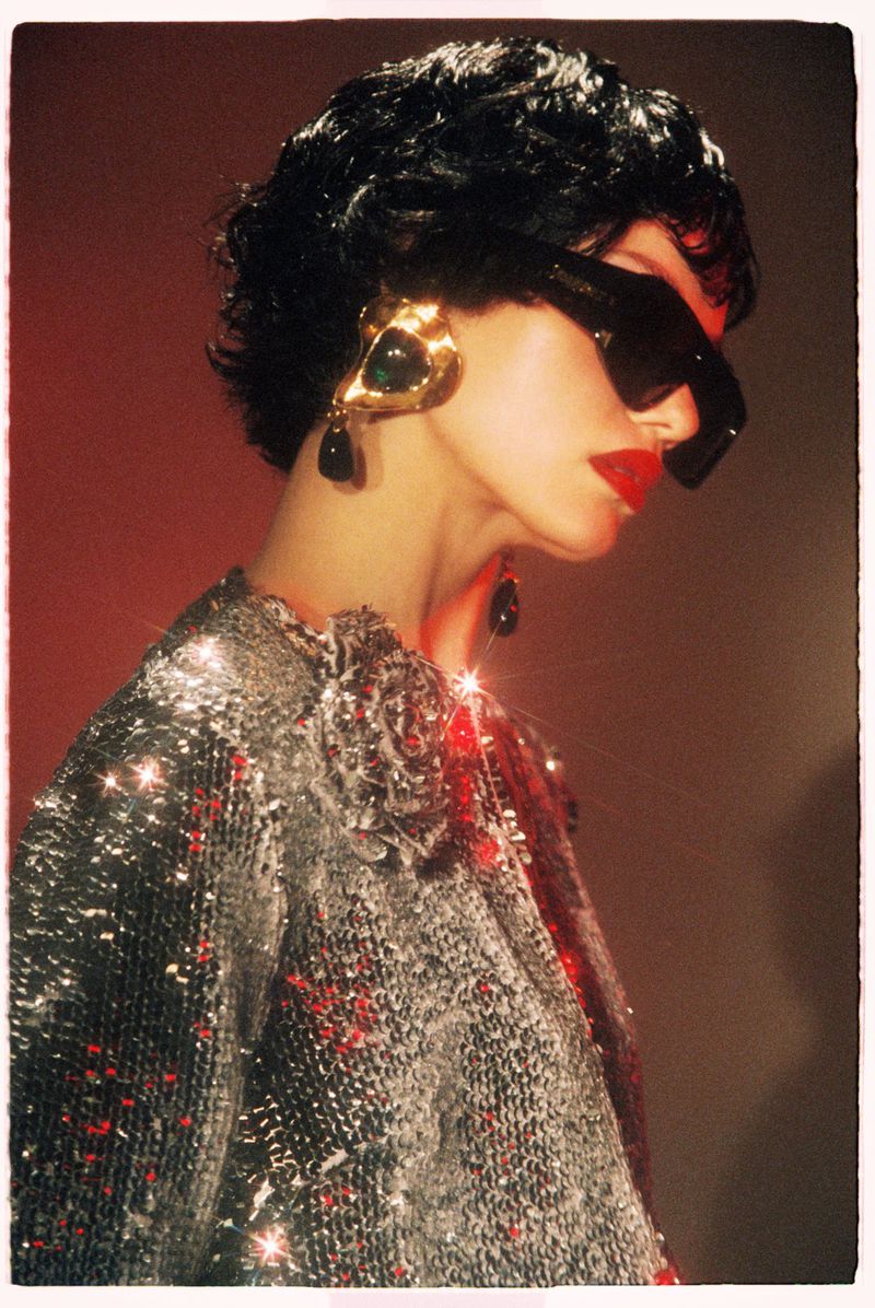Model wearing dark sunglasses, gold earrings, and a sparkling silver sequin top against a warm gradient background.