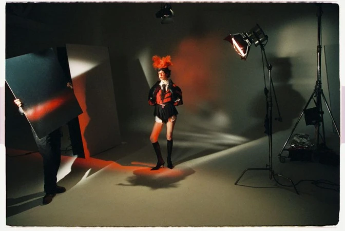 Behind-the-scenes fashion studio shot showing a model in a theatrical red and black outfit with feathered headpiece posing amidst lighting equipment