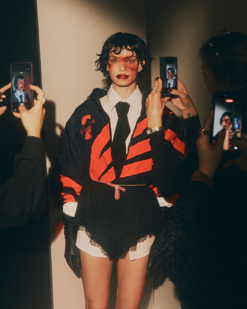 Model surrounded by smartphones recording video, wearing black and red jacket, white shirt, dark tie, and gloves