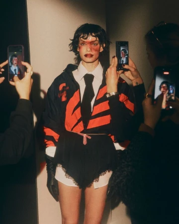 Model surrounded by smartphones recording video, wearing black and red jacket, white shirt, dark tie, and gloves