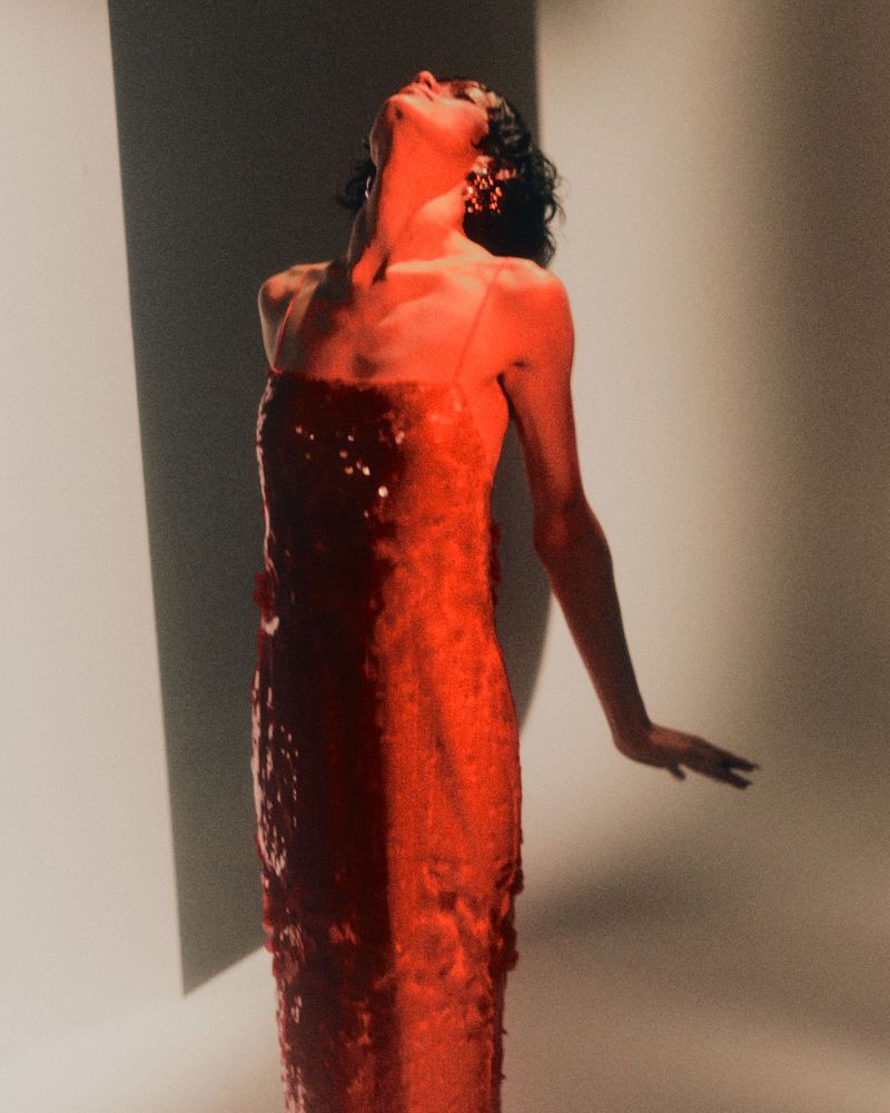 Model in a red sequin dress with head tilted back against a bright white studio background under warm dramatic red lighting