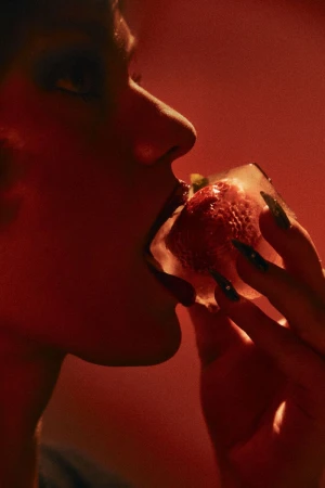 Close-up profile of a woman holding an ice cube containing a strawberry near her mouth under warm red lighting