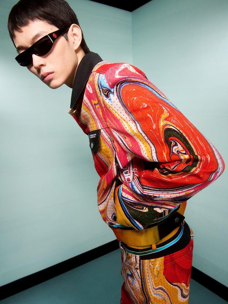Asian male fashion model wearing a colorful, patterned jacket and sunglasses poses against a light blue wall in a studio setting.