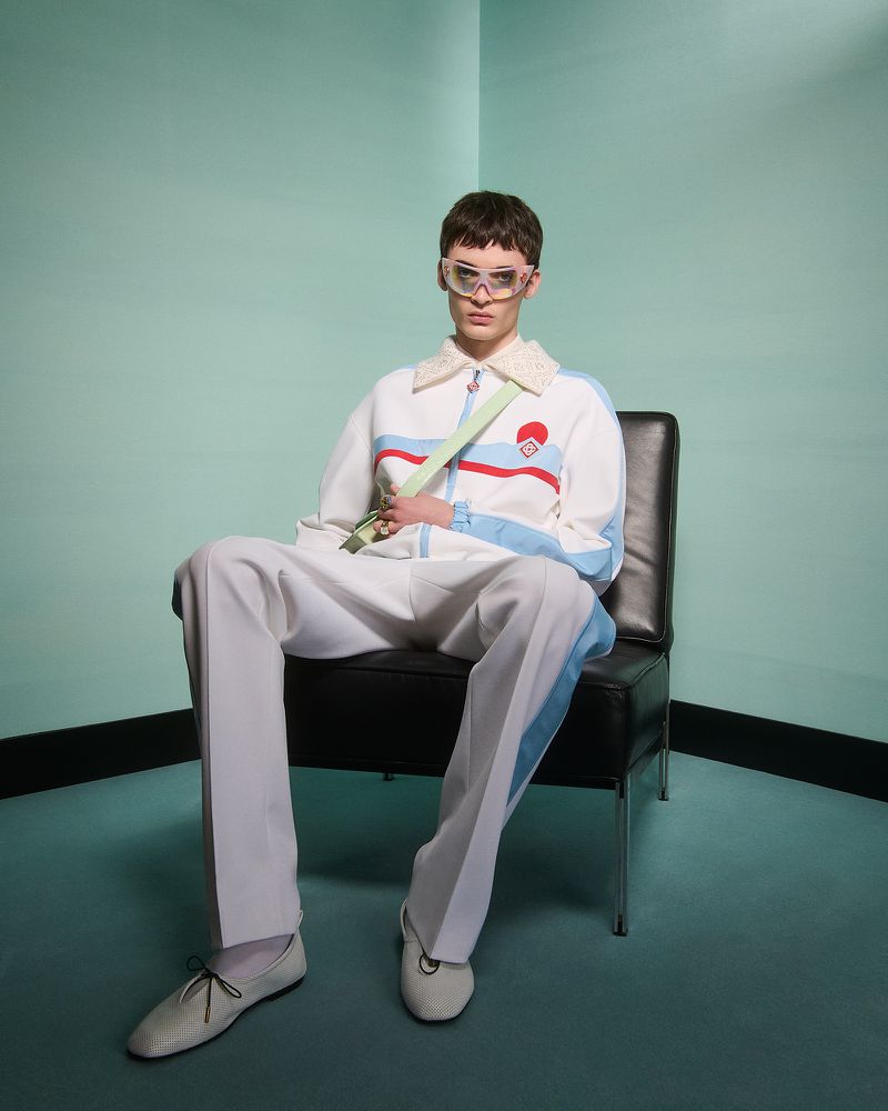 A person sits on a black chair against a teal background wearing a white track suit with blue stripes and clear eyewear.