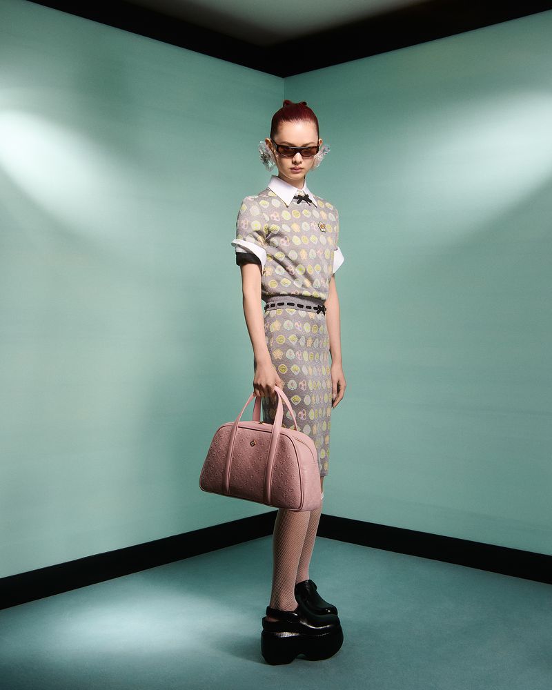 A woman poses in a grey patterned skirt and top with a pink purse and black platform shoes, inside a mint green corner set.