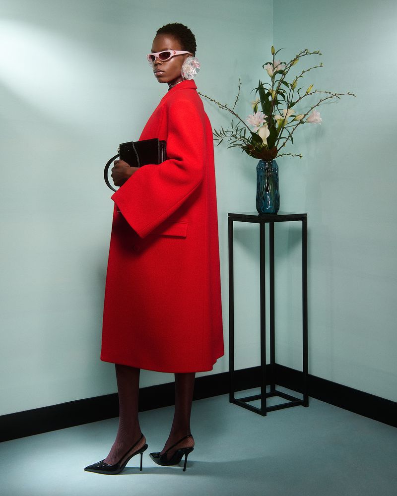 Fashion shot of a Black woman in a red coat and black heels, holding a black bag, with a vase of white flowers in the background.