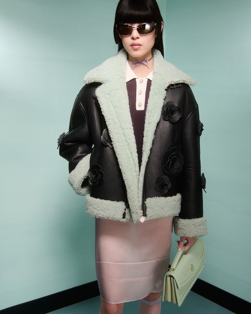 A stylish woman in sunglasses models a black shearling coat with floral appliqués, holding a mint green bag in a fashion setting.
