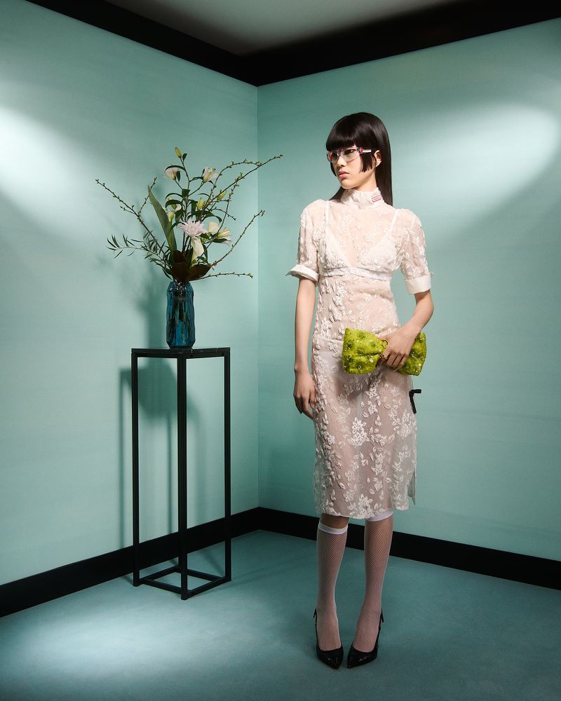 A woman in a white floral dress and heels poses in front of a light blue wall next to a vase of white flowers on a black stand.