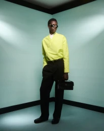 Model in lime green shirt and black trousers holding a handbag against a teal corner set for Casablanca AW 2025.