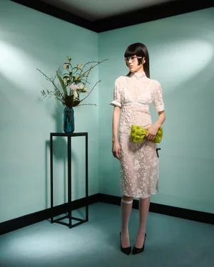 Model in sheer lace dress holding a lime green clutch standing in a mint green room with a floral vase on a metal stand.