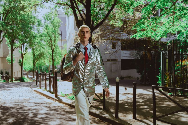 Young man with dyed blonde hair wearing a metallic silver jacket and red tie walking down an urban street.