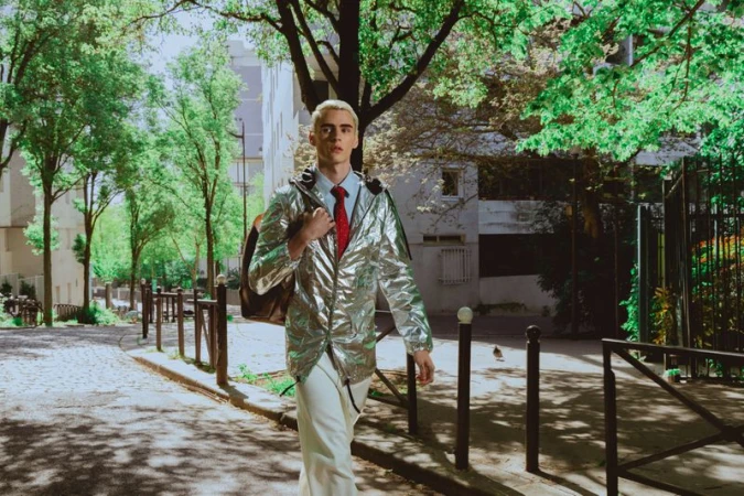 Young man with dyed blonde hair wearing a metallic silver jacket and red tie walking down an urban street.