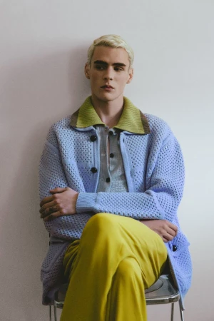 Blonde male model wearing a light blue knit cardigan with a lime collar and yellow trousers seated against a plain wall.
