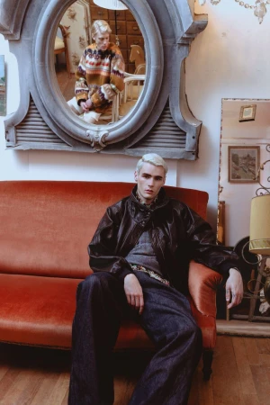 A male model seated on a red velvet couch wearing a dark leather jacket, with his reflection appearing in a large ornate mirror above.