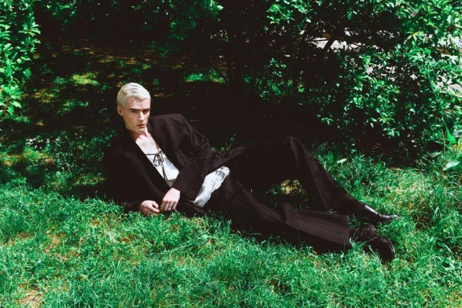 Model with bleached blonde hair lying in green grass wearing a dark suit jacket and trousers for Vanity Teen Magazine.
