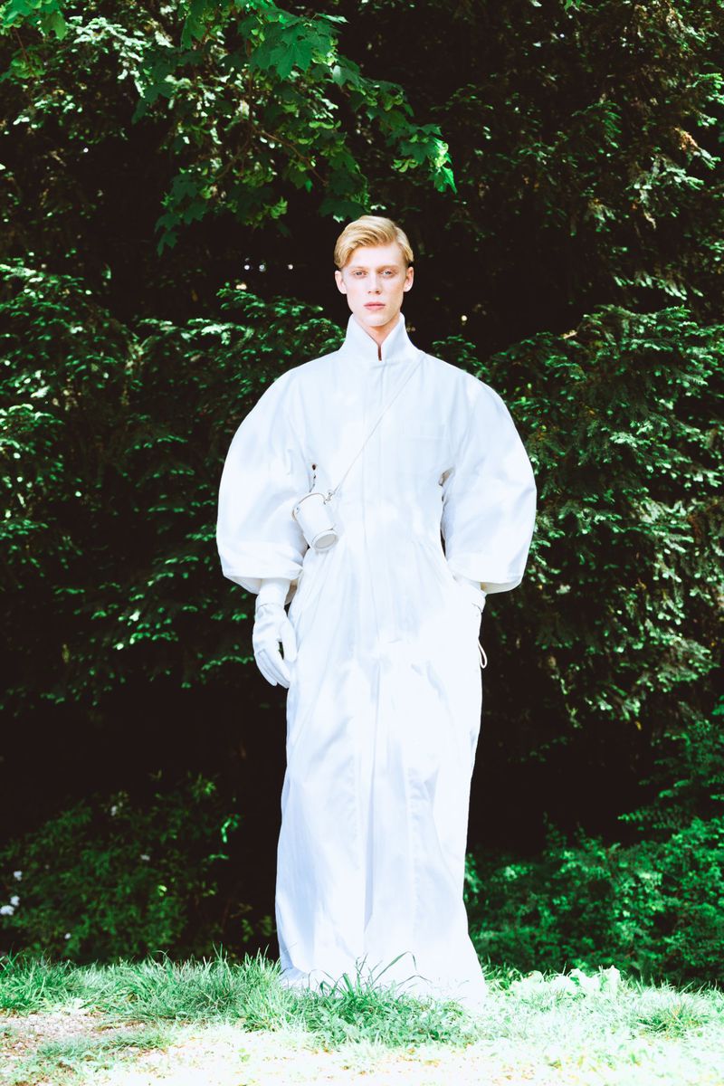 Model in a voluminous white jumpsuit standing against a lush green foliage background