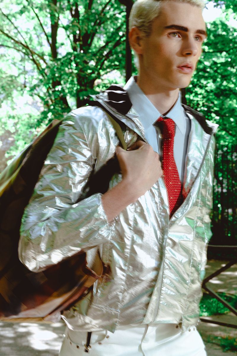 Young male model with blonde hair wearing a metallic silver jacket, white shirt, and red tie outdoors near trees