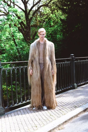 Model standing on a stone bridge wearing a long fringed coat against a lush green forest background