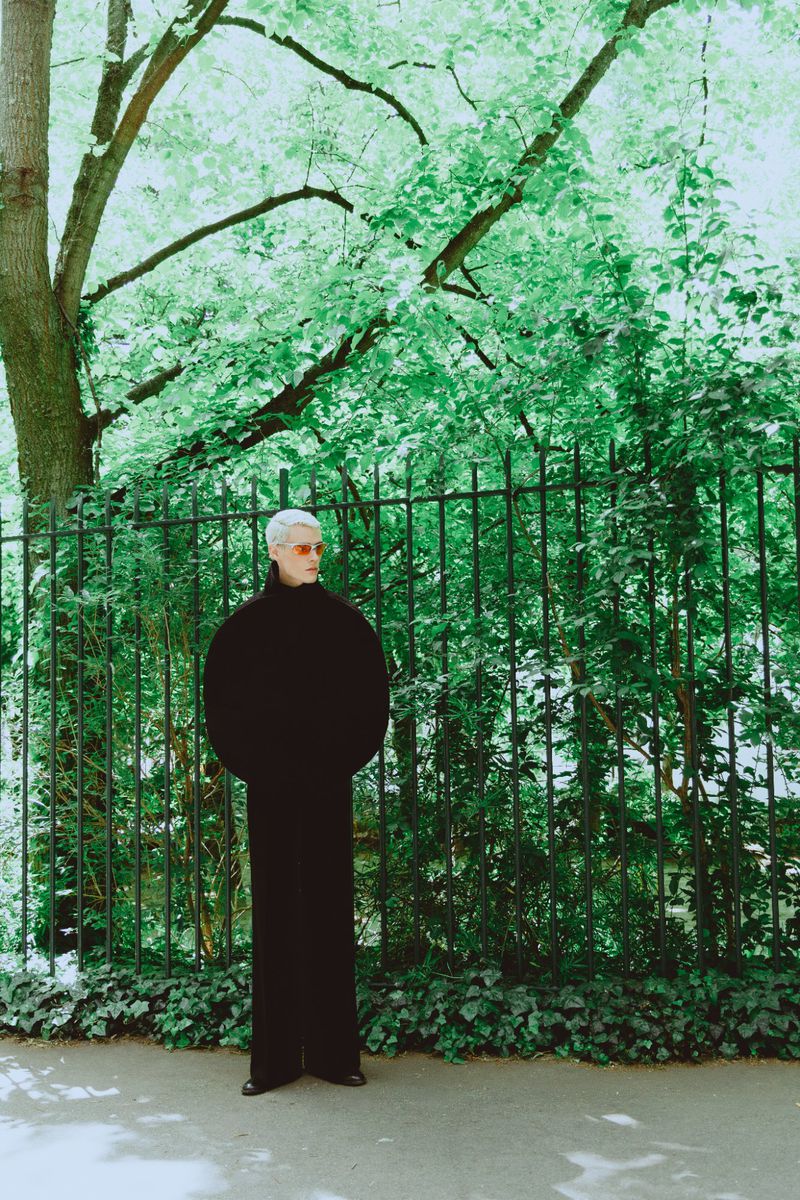 Model wearing dark voluminous sweater standing before a dark metal fence and green trees for Vanity Teen Magazine.