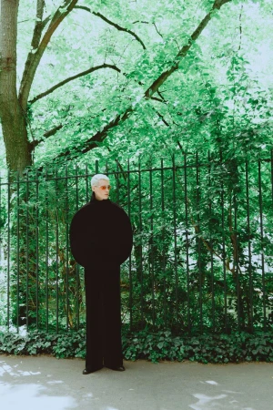 Model wearing dark voluminous sweater standing before a dark metal fence and green trees for Vanity Teen Magazine.