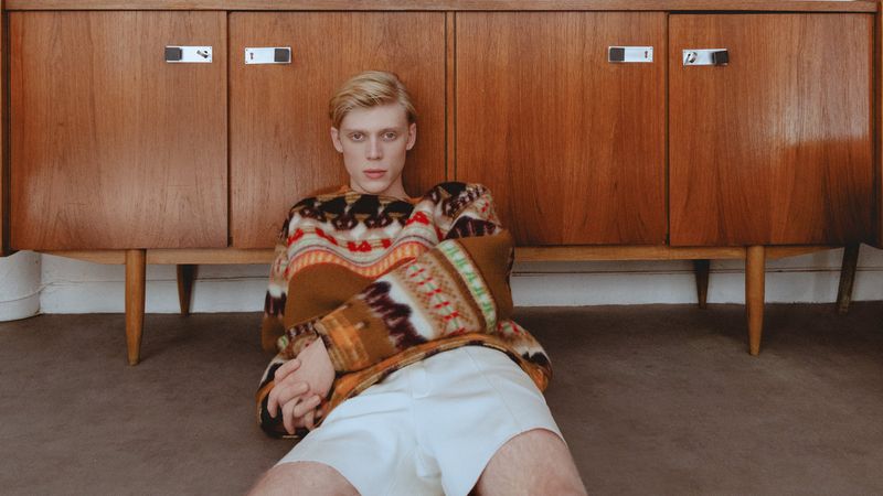 Blonde male model wearing a patterned sweater seated on the floor against a wooden sideboard for Vanity Teen Magazine.