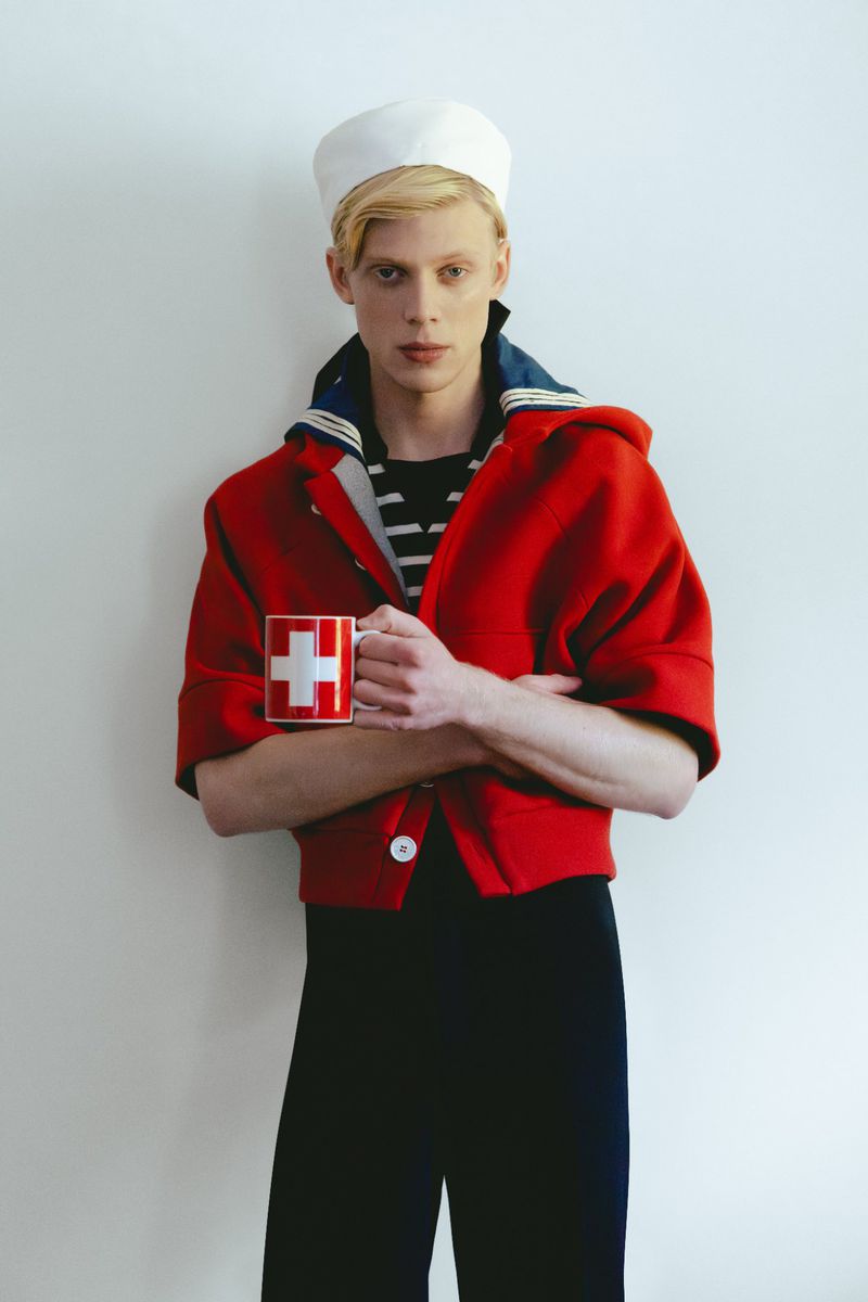 Blonde male model wearing a red jacket and white sailor cap holding a red mug with a white cross against a white wall.