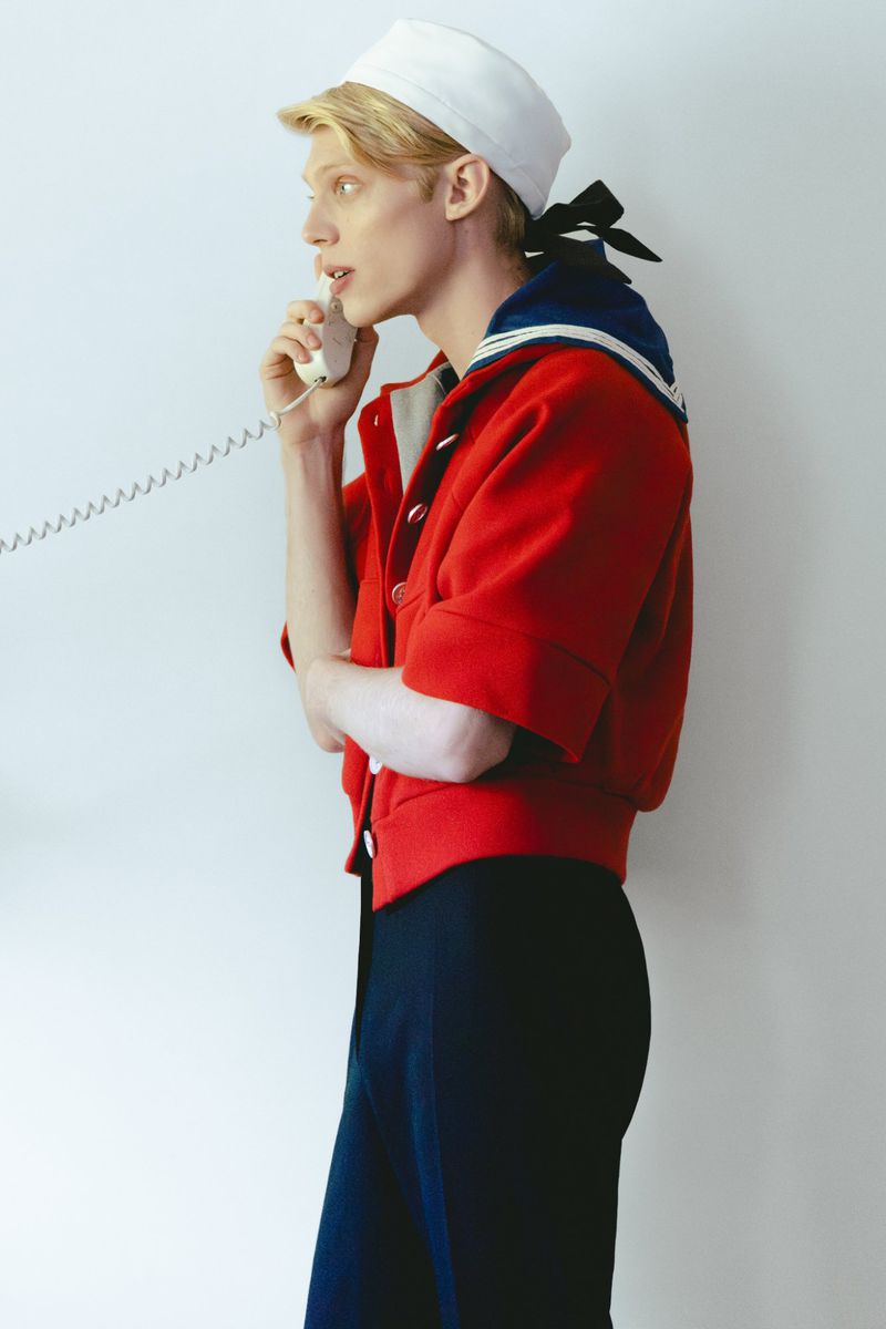 Model wearing a sailor-style hat and red jacket holding a white rotary telephone receiver against a plain white background.