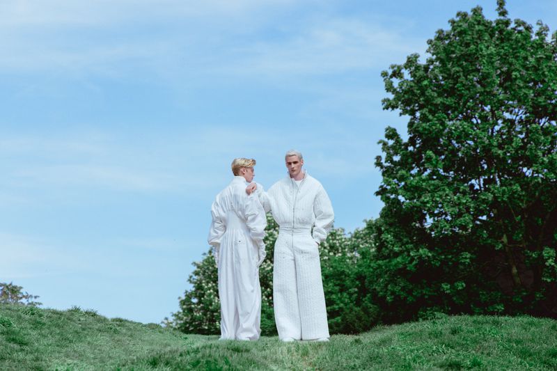 Two male models in matching white textured outfits standing on a grassy hill under a bright sky with trees.