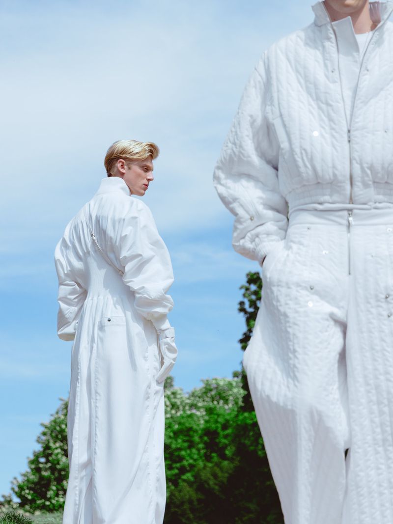 Two people wearing white outfits stand outdoors against a blue sky and trees in this fashion-forward editorial style photograph.