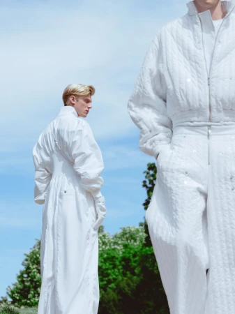 Two people wearing white outfits stand outdoors against a blue sky and trees in this fashion-forward editorial style photograph.